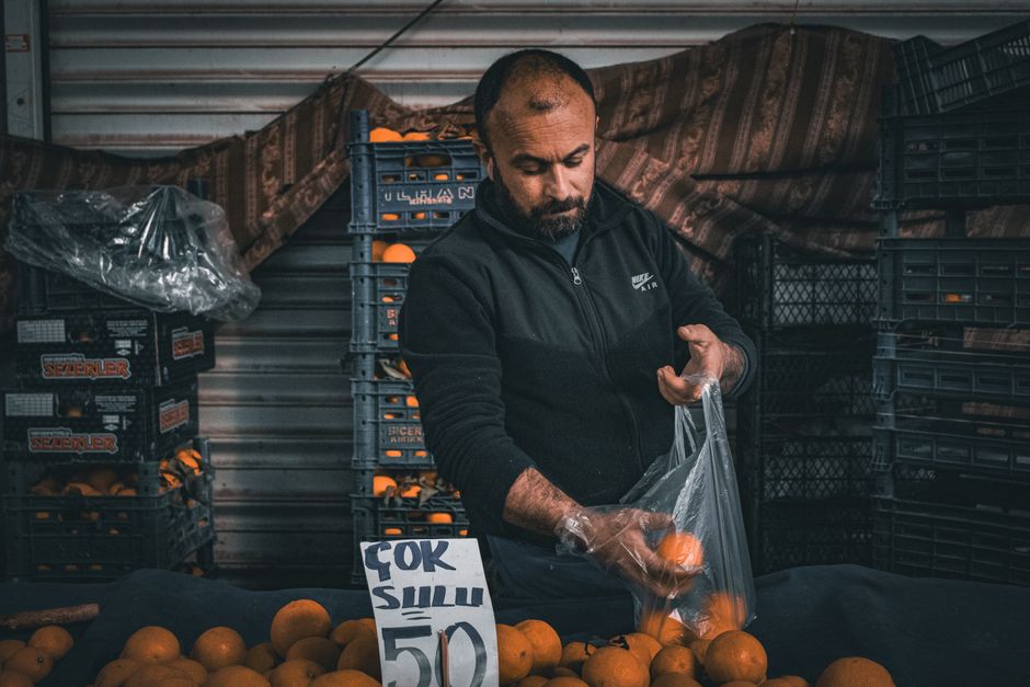 A man wearing a black Nike Air zip-up jacket and transparent gloves is packaging fresh oranges into a plastic bag at a market stall. Behind him, there are stacked crates filled with oranges and a meta