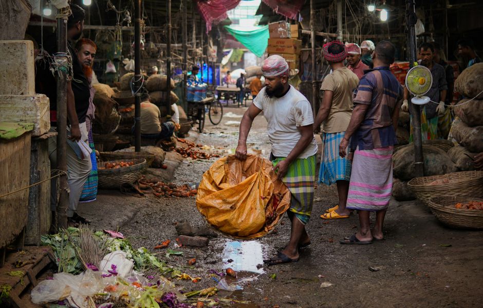 A man wearing a black Nike Air zip-up jacket and transparent gloves is packaging fresh oranges into a plastic bag at a market stall. Behind him, there are stacked crates filled with oranges and a meta